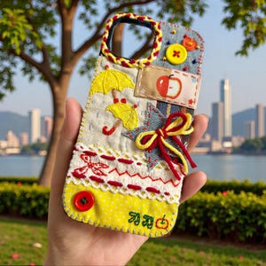 Puede incluir: Una funda de teléfono hecha a mano con un diseño de patchwork. Presenta paraguas amarillos bordados, una manzana roja y un lazo amarillo y rojo. La funda tiene una base de tela vaquera y amarilla, con costuras y botones rojos y amarillos.