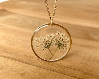 Könnte beinhalten: Eine goldene Kettenkette mit einem runden Anhänger, in dem eine gepresste weiße Blume in klarem Harz eingebettet ist.
