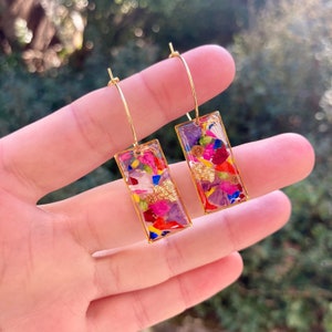 Op de afbeelding: Gouden oorringen met rechthoekige hangers. De hangers zijn gevuld met kleurrijke gedroogde bloemen en bladgoud. De oorbellen worden in een hand gehouden tegen een vage groene achtergrond.