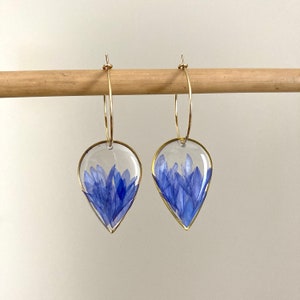 Può includere: Un paio di orecchini a cerchio dorati con ciondoli in resina a forma di goccia. Ogni ciondolo presenta un gruppo di fiori blu pressati.