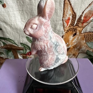 May include: A pink and gray carved stone rabbit figurine. The bunny is sitting on a clear, round display stand with a black base. The background features a floral patterned fabric.