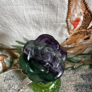 May include: A green and purple fluorite rose carved from a natural crystal. The rose is sitting on a green glass base.