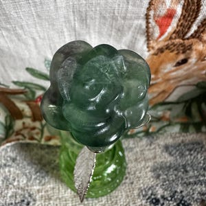 May include: A green fluorite rose carved from a single piece of crystal. The rose is sitting on a green glass bottle with a silver leaf.