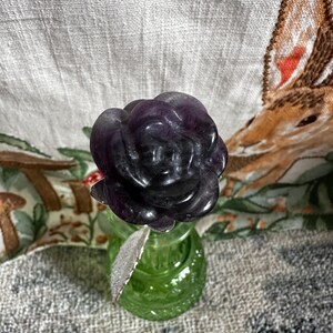 May include: A purple amethyst rose carved from a single stone, sitting in a green glass vase.