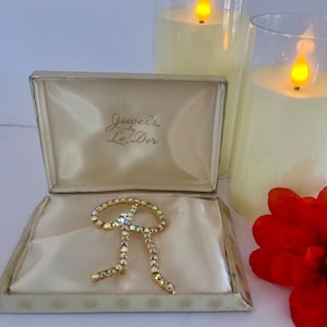 May include: A vintage bow-shaped brooch with iridescent rhinestones, presented in a cream-colored jewelry box. The box is open, revealing the brooch on a satin lining. The box is labeled "Jewels Le' Dor".