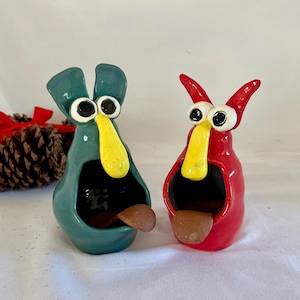 May include: Two whimsical ceramic figurines, one teal and one red, each with large eyes, a yellow beak, and a brown tongue. The teal figurine has two upright ear-like protrusions. The red figurine has two curved horns. Both have open mouths.