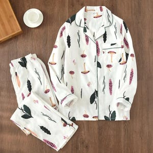 Può includere: Set pigiama bianco con stampa botanica con funghi, foglie e forme a piuma in rosa, nero e verde. La parte superiore a maniche lunghe ha un colletto e finiture verdi. I pantaloni abbinati sono piegati.