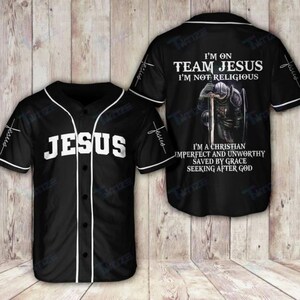 Può includere: Maglia da baseball nera con finiture bianche e la scritta "JESUS" sul davanti. Le maniche hanno la parola "Jesus" e una croce. Il retro della maglia presenta la scritta "I'M ON TEAM JESUS I'M NOT RELIGIOUS" e una grafica di un cavaliere in ginocchio.
