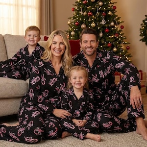 May include: A family of four wearing matching black pajamas with pink bow patterns. They are sitting in front of a decorated Christmas tree. The pajamas have a button-down top and pants.