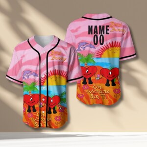Könnte beinhalten: Rosa Baseball-Trikot mit tropischem Design mit Delfinen, Palmen und herzförmigen Figuren. Das Trikot hat schwarze Verzierungen und den Text "Bad Bunny" auf der Vorder- und Rückseite. Auf der Rückseite steht außerdem der Text "NAME 00" und "Un Verano Sin Ti".
