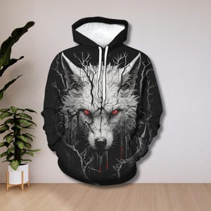 May include: Black hoodie with a striking wolf design. The wolf's face is depicted with red eyes and a white and grey fur pattern, framed by tree-like branches. The hood has a white inner lining. A unique piece of clothing.