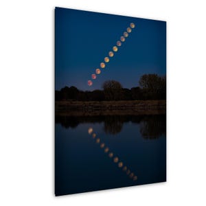 May include: A photograph of the moon's phases in a diagonal line across a dark blue sky. The moon's reflection is visible in the water below, with trees and a shoreline in the background.