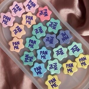 Puede incluir: Una colección de objetos en forma de estrella, de colores pastel, con el texto "PAN ME" en azul oscuro. Los objetos están dispuestos en un recipiente transparente, con colores como rosa, amarillo, verde y azul. La estética general es suave y juguetona.
