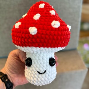 May include: A handmade, plush mushroom toy with a red cap dotted with white spots. The stem is white, and the mushroom has a smiling face with black eyes. The toy is made of soft, textured yarn.