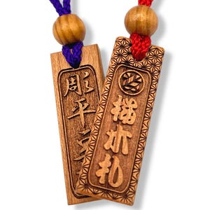 May include: Two wooden Japanese omamori charms with carved characters and decorative borders. One charm has purple cord, the other red. Both have wooden beads at the top. The charms are rectangular and feature detailed inscriptions.