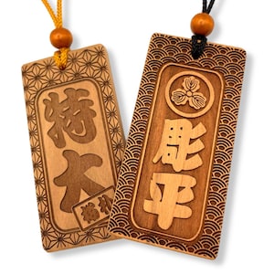 May include: Two wooden Japanese omamori charms. One charm has Japanese characters and a small rectangular tag. The other charm has Japanese characters and a floral emblem. Both charms have intricate border designs and are suspended by cords.