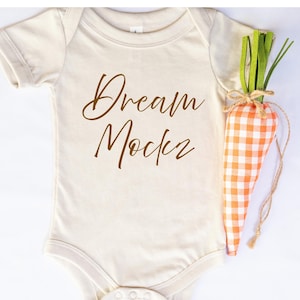 May include: A cream-coloured baby onesie with the words "Dream Maker" in brown script. A decorative carrot made of orange and white checkered fabric and green felt tops is next to the onesie.