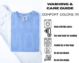 May include: A light blue Comfort Colors 1717 t-shirt is folded on top of a pair of light-wash jeans. The shirt has a crew neck and a small tag. Washing and care instructions are listed on the right.