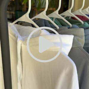 May include: A selection of hooded sweatshirts hanging on white coat hangers. The garments are in a range of colours, including white, grey, green, and blue. The hoodies are displayed on a clothes rail, with a blurred background of foliage.