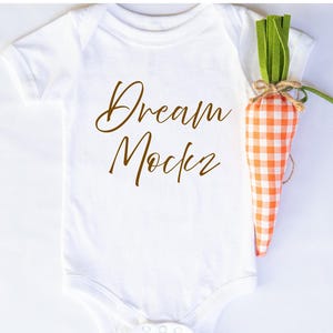 May include: White baby bodysuit with the words "Dream Maker" in brown script. A decorative carrot made of orange and white gingham fabric with green felt leaves is next to the bodysuit.