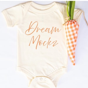 May include: Cream-coloured baby onesie with the words "Dream Maker" in a copper-coloured script. A decorative carrot made of orange and white checkered fabric and green fabric is next to the onesie.