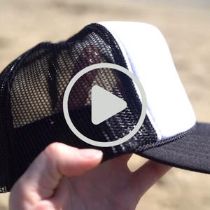 Könnte beinhalten: Eine schwarz-weiße Trucker-Mütze mit Mesh-Rückseite und gebogenem Schirm. Die Vorderseite ist weiß und der Schirm ist schwarz. Die Mütze wird vor einem sandigen Hintergrund gehalten.