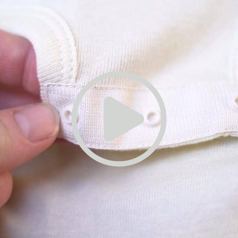 May include: Close-up of a cream-colored onesie with snap closures. The fabric appears to be soft and knitted. The snap closures are white and round, providing a secure fastening for the garment. The onesie is likely for a baby or toddler.