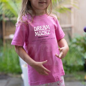 Puede incluir: Una camiseta rosa con el texto blanco "DREAM MOCKZ", las palabras "MOCKUP STUDIO" y texto adicional. La camiseta de manga corta está siendo usada. La imagen es de un niño.