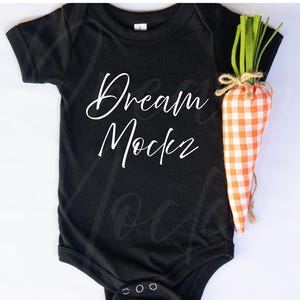 May include: Black baby bodysuit with the text "Dream Mockz" in white script. A decorative carrot made of orange and white gingham fabric with green fabric leaves is next to the bodysuit.