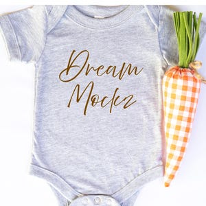 May include: A grey baby bodysuit with the words "Dream Maker" in brown script. A decorative carrot with an orange and white gingham pattern is next to the bodysuit. The carrot has green fabric for the stem.