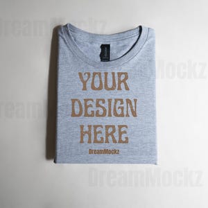 以下が含まれることがあります： 折りたたまれたライトグレーのTシャツ。茶色のヴィンテージ風フォントで「YOUR DESIGN HERE」と書かれています。ブランドタグが首元に見えます。「DreamMockz」の文字が下に印刷されています。