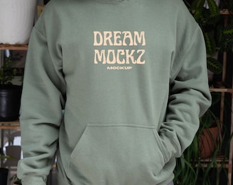 May include: Sage green hoodie with a front pocket and a drawstring hood. The text "DREAM MOCKZ MOCKUP" is printed on the front in a light tan color. The hoodie is made of a soft material.