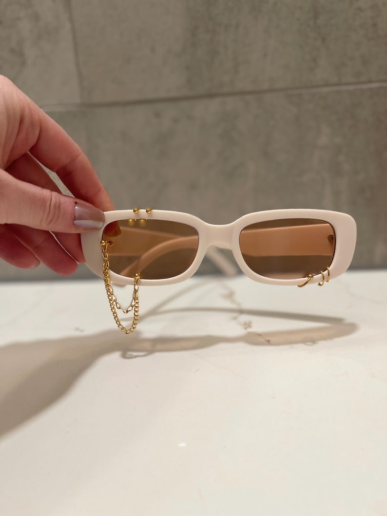 Op de afbeelding: Off-white zonnebril met een rechthoekig montuur en lichtbruine glazen. De zonnebril heeft goudkleurige ketting- en hoepelaccenten. De zonnebril wordt door een hand vastgehouden.
