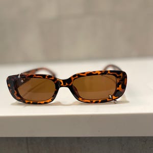 Op de afbeelding: Rechthoekige zonnebril met een montuur met schildpadpatroon en bruine glazen. Deze zonnebril heeft een klassiek ontwerp met een moderne twist, geschikt voor verschillende gezichtsvormen. Het montuur heeft een gespikkeld patroon in bruin- en zwarttinten.