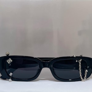 Op de afbeelding: Zwarte zonnebril met een rechthoekig montuur. De zonnebril is versierd met zilverkleurige studs en een ketting. De glazen zijn donker en het montuur is dik en glanzend.