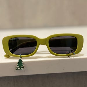 Op de afbeelding: Limoengroene zonnebril met donkere glazen. De monturen hebben kleine decoratieve piercings en een bungelende groene boomvormige bedel met het woord "FRESH". De zonnebril heeft een rechthoekige vorm en een modern design.