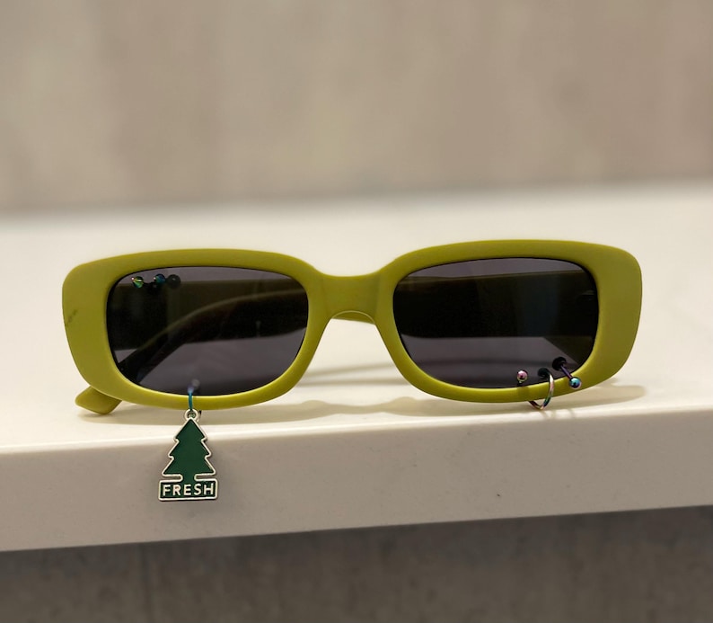 Op de afbeelding: Limoengroene zonnebril met donkere glazen. De monturen hebben kleine decoratieve piercings en een bungelende groene boomvormige bedel met het woord "FRESH". De zonnebril heeft een rechthoekige vorm en een modern design.