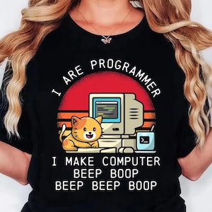 Op de afbeelding: Zwart T-shirt met een afbeelding van een retro computer, een lachende oranje kat en de tekst "I ARE PROGRAMMER. I MAKE COMPUTER BEEP BOOP BEEP BEEP BOOP" in het wit. Het ontwerp bevat een rode en oranje zonsondergang.