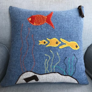 Puede incluir: Una almohada de denim azul con peces y algas bordadas. Los peces son naranja y amarillos. La almohada tiene un tirador de cremallera de cuero marrón.