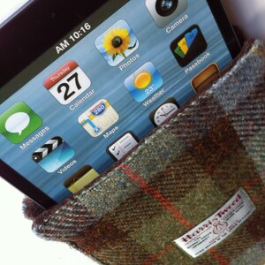 Może przedstawiać: Zbliżenie na ekran smartfona, na kt&oacute;rym widoczne są r&oacute;żne ikony aplikacji, w tym Wiadomości, Kalendarz, Zdjęcia, Pogoda i Passbook. Telefon jest częściowo zakryty brązową i zieloną tkaniną tweedową z nadrukiem "Harris Tweed".