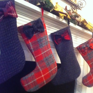 May include: Four Christmas stockings hanging on a white mantel. The stockings are made of a plaid fabric with a red and gray pattern. Each stocking has a bow made of a different fabric. The bows are red, black, red, and black.