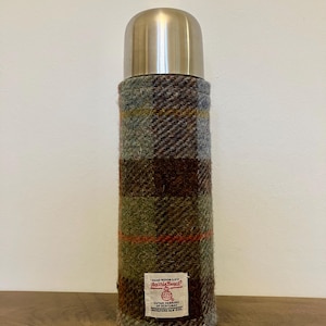 Może przedstawiać: Termos ze stali nierdzewnej ze srebrną pokrywką i pokrowcem z tkaniny Harris Tweed. Pokrowiec ma wz&oacute;r w kratę w odcieniach brązu, zieleni i szarości, z małą etykietą. Idealny do utrzymywania temperatury napoj&oacute;w.