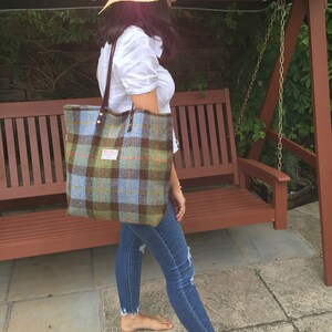 May include: A large tote bag with a plaid pattern in shades of green, blue, and brown. The bag has brown leather handles and a label. The person is wearing a straw hat, white shirt, and blue jeans.