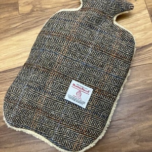 Puede incluir: Una botella de agua caliente de tweed marrón y negro con forro de vellón blanco. La botella tiene una etiqueta que dice "Harris Tweed" con una pequeña imagen de una oveja y una rueca.