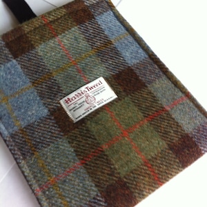 Op de afbeelding: Een bruine, groene en blauwe geruite stoffen tas met een wit label waarop "Harris Tweed" staat en het Harris Tweed Orb-logo. Het label bevat ook de tekst "The Harris Tweed Certification Mark" en "HAND WOVEN IN THE OUTER HEBRIDES".