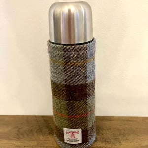 Może przedstawiać: Termos ze stali nierdzewnej z poszewką z tkaniny w kratę Harris Tweed w odcieniach szarości, brązu i zieleni. Termos ma za zadanie utrzymywać napoje gorące lub zimne. Etykieta głosi "Harris Tweed".