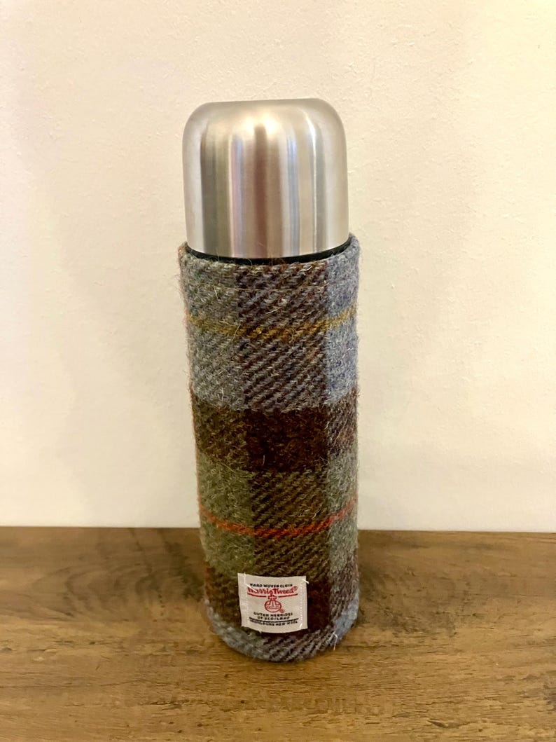 Może przedstawiać: Termos ze stali nierdzewnej z poszewką z tkaniny w kratę Harris Tweed w odcieniach szarości, brązu i zieleni. Termos ma za zadanie utrzymywać napoje gorące lub zimne. Etykieta głosi "Harris Tweed".