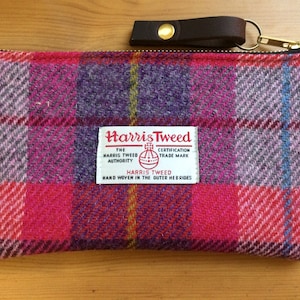 Puede incluir: Una bolsa de cremallera de color rosa, morado y gris a cuadros con una correa de cuero marrón y un tirador de cremallera de metal dorado. La bolsa tiene una etiqueta blanca con el texto "Harris Tweed" y el logotipo de Harris Tweed Orb.