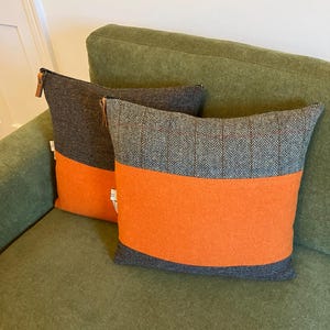 May include: Two decorative throw pillows with a gray and orange color scheme. The pillows have a tweed-like fabric with a herringbone pattern. The pillows are on a green couch.