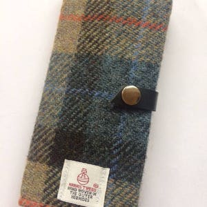 May include: A blue, green, and orange plaid wallet with a black leather strap and a gold button. The wallet is made of Harris Tweed, handwoven in the Outer Hebrides.